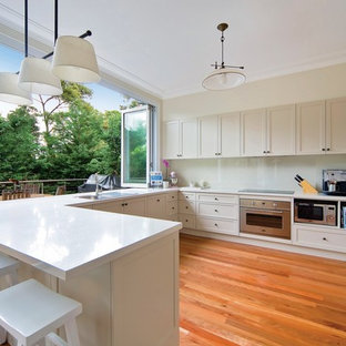 This is an example of a transitional u-shaped eat-in kitchen in Sydney with white cabinets, white splashback, stainless steel appliances, medium hardwood floors, white benchtop, a drop-in sink, shaker cabinets, glass sheet splashback, a peninsula and red floor.