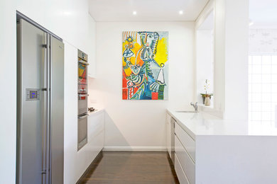 This is an example of a contemporary galley open plan kitchen in Sydney with a submerged sink, flat-panel cabinets, white cabinets, engineered stone countertops, glass sheet splashback and stainless steel appliances.