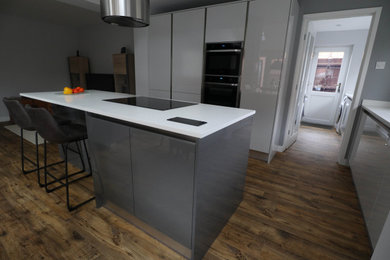 Medium sized contemporary open plan kitchen in Cambridgeshire with a submerged sink, flat-panel cabinets, grey cabinets, composite countertops, black appliances, cement flooring, an island, brown floors and white worktops.