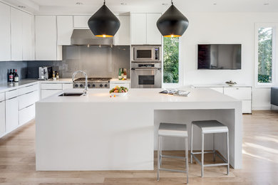 Photo of a medium sized contemporary l-shaped open plan kitchen in Seattle with a built-in sink, flat-panel cabinets, white cabinets, grey splashback, stainless steel appliances, light hardwood flooring and an island.