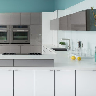Example of a trendy u-shaped kitchen design in Newark with a drop-in sink, flat-panel cabinets, white cabinets, white backsplash, glass sheet backsplash, stainless steel appliances and a peninsula