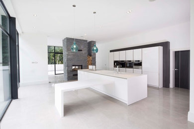 Идея дизайна: огромная параллельная кухня-гостиная в стиле модернизм с монолитной мойкой, плоскими фасадами, белыми фасадами, столешницей из акрилового камня, черной техникой, полом из керамогранита и островом