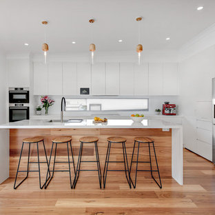 This is an example of a large modern l-shaped kitchen in Sydney with an undermount sink, flat-panel cabinets, white cabinets, quartz benchtops, white splashback, glass sheet splashback, stainless steel appliances, with island, brown floor, white benchtop and medium hardwood floors.