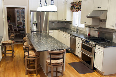 This is an example of a traditional u-shaped kitchen in Cincinnati with a double-bowl sink, raised-panel cabinets, white cabinets, granite worktops, grey splashback, glass tiled splashback, stainless steel appliances, light hardwood flooring and an island.