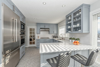 Mid-sized transitional l-shaped ceramic tile and multicolored floor enclosed kitchen photo in Vancouver with a double-bowl sink, shaker cabinets, blue cabinets, solid surface countertops, white backsplash, subway tile backsplash, stainless steel appliances and a peninsula