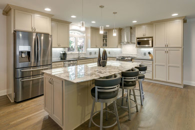 Example of a large minimalist l-shaped medium tone wood floor and brown floor open concept kitchen design with an undermount sink, recessed-panel cabinets, beige cabinets, quartz countertops, beige backsplash, mosaic tile backsplash, stainless steel appliances, an island and black countertops