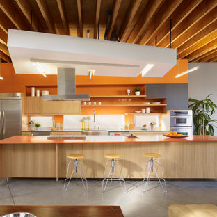 Diseño de cocina de galera, actual, con armarios con paneles lisos, puertas de armario de madera clara, salpicadero naranja, electrodomésticos de acero inoxidable, suelo de cemento, una isla, suelo gris y encimeras naranjas