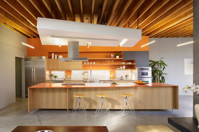 Diseño de cocina actual con armarios con paneles lisos, puertas de armario de madera clara, salpicadero naranja, electrodomésticos de acero inoxidable, suelo de cemento, una isla, suelo gris y encimeras naranjas