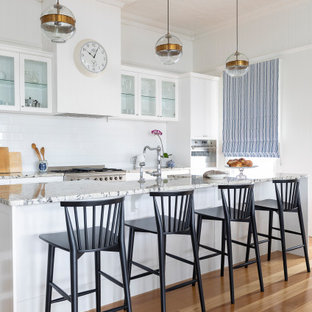 This is an example of a mid-sized transitional galley open plan kitchen in Brisbane with a farmhouse sink, recessed-panel cabinets, white cabinets, granite benchtops, cement tile splashback, stainless steel appliances, light hardwood floors, with island, grey benchtop, white splashback and brown floor.