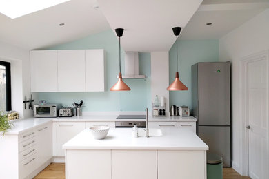 Medium sized contemporary l-shaped open plan kitchen in London with flat-panel cabinets, white cabinets, composite countertops, glass sheet splashback, white appliances, light hardwood flooring, an island, an integrated sink, blue splashback and beige floors.
