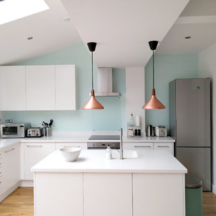 Mid-sized contemporary l-shaped open plan kitchen in London with flat-panel cabinets, white cabinets, solid surface benchtops, glass sheet splashback, white appliances, light hardwood floors, with island, an integrated sink, blue splashback and beige floor.