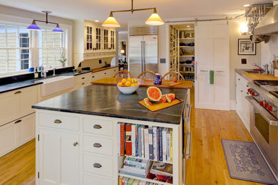 Example of a large cottage u-shaped light wood floor kitchen pantry design in Portland Maine with a farmhouse sink, white cabinets, white backsplash, ceramic backsplash, stainless steel appliances, an island, shaker cabinets and soapstone countertops