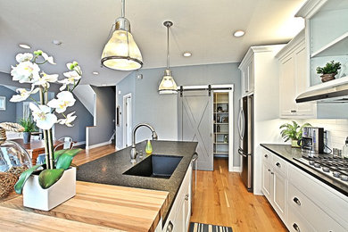 Example of a beach style single-wall medium tone wood floor open concept kitchen design in Grand Rapids with an undermount sink, shaker cabinets, white cabinets, white backsplash, ceramic backsplash, stainless steel appliances and an island