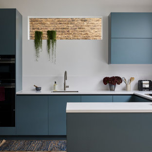 Réalisation d'une cuisine ouverte design en U avec un placard à porte plane, des portes de placard bleues, un plan de travail en surface solide, une crédence blanche, une crédence en quartz modifié, un sol en bois brun, un plan de travail blanc, un évier encastré, un électroménager noir, une péninsule et un sol marron.