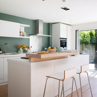 This is an example of a large contemporary u-shaped open plan kitchen in London with an integrated sink, flat-panel cabinets, white cabinets, laminate benchtops, green splashback, stainless steel appliances, light hardwood floors, with island, beige floor and white benchtop.