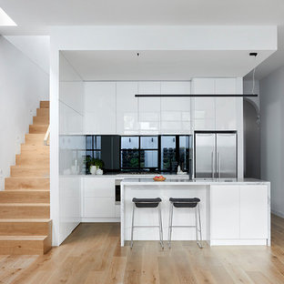 This is an example of a contemporary open plan kitchen in Melbourne with white cabinets, marble benchtops, black splashback, glass sheet splashback, stainless steel appliances, with island, white benchtop, flat-panel cabinets, light hardwood floors and beige floor.