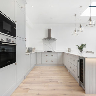 This is an example of a large classic grey and white u-shaped open plan kitchen in London with a submerged sink, shaker cabinets, grey cabinets, grey splashback, stainless steel appliances, light hardwood flooring, a breakfast bar, beige floors and white worktops.