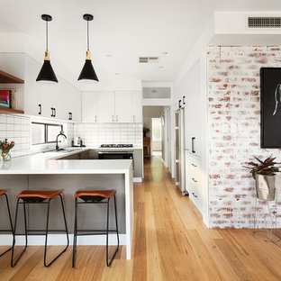 Photo of a mid-sized contemporary u-shaped kitchen in Melbourne with flat-panel cabinets, white cabinets, white splashback, ceramic splashback, medium hardwood floors, brown floor, white benchtop, a drop-in sink, panelled appliances and a peninsula.