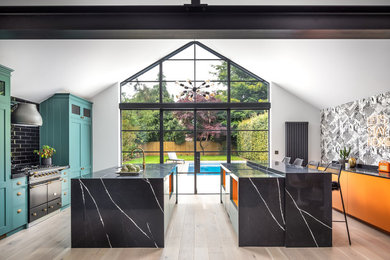 Photo of a contemporary galley open plan kitchen in Kent with multiple islands, shaker cabinets, blue cabinets, black splashback, black appliances, light hardwood flooring, beige floors and black worktops.