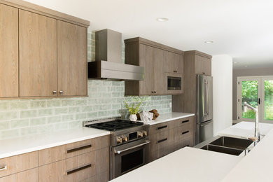 Example of a large minimalist galley kitchen design in Minneapolis with a farmhouse sink, flat-panel cabinets, light wood cabinets, solid surface countertops, green backsplash, subway tile backsplash, stainless steel appliances and an island