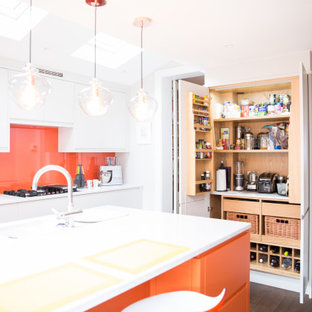 Offene, Mittelgroße Moderne Küche in U-Form mit Waschbecken, flächenbündigen Schrankfronten, orangefarbenen Schränken, Mineralwerkstoff-Arbeitsplatte, Küchenrückwand in Orange, Rückwand aus Glasfliesen, dunklem Holzboden, Kücheninsel, braunem Boden und weißer Arbeitsplatte in London