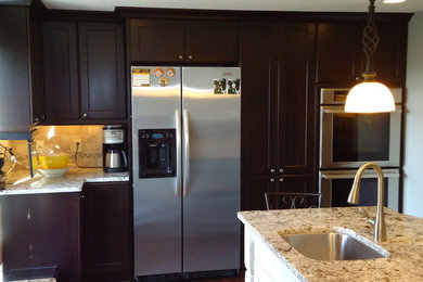 Example of a large l-shaped medium tone wood floor kitchen pantry design in Other with dark wood cabinets, granite countertops, stainless steel appliances, an island, an undermount sink, recessed-panel cabinets, beige backsplash and stone tile backsplash