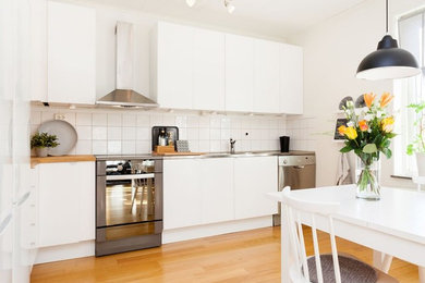 This is an example of a contemporary single-wall kitchen/diner in Other with an integrated sink, flat-panel cabinets, white cabinets, stainless steel worktops, white splashback, stainless steel appliances, medium hardwood flooring, brown floors and grey worktops.