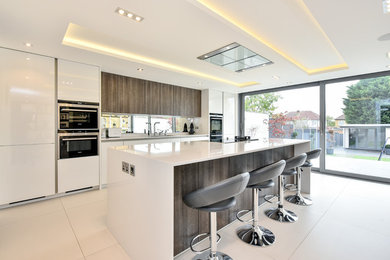 This is an example of a medium sized modern single-wall open plan kitchen in London with a built-in sink, flat-panel cabinets, white cabinets, quartz worktops, metallic splashback, glass sheet splashback, black appliances, ceramic flooring, an island and white floors.
