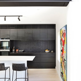 Photo of a contemporary galley kitchen in Sydney with flat-panel cabinets, black cabinets, black splashback, stainless steel appliances, light hardwood floors, with island, beige floor and black benchtop.