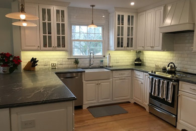 Example of a mid-sized classic u-shaped light wood floor and brown floor eat-in kitchen design in Philadelphia with a farmhouse sink, raised-panel cabinets, white cabinets, granite countertops, white backsplash, porcelain backsplash, stainless steel appliances, an island and gray countertops