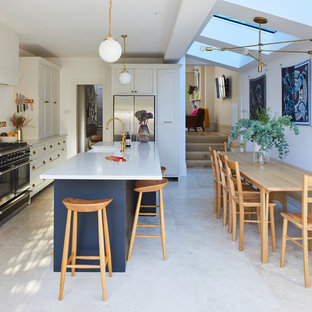 Design ideas for a classic l-shaped kitchen/diner in London with a belfast sink, shaker cabinets, white cabinets, white splashback, stainless steel appliances, an island, beige floors and white worktops.