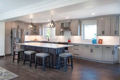 Photo of a large contemporary l-shaped kitchen/diner in Toronto with a belfast sink, flat-panel cabinets, grey cabinets, engineered stone countertops, white splashback, ceramic splashback, stainless steel appliances, medium hardwood flooring, an island, brown floors and white worktops.