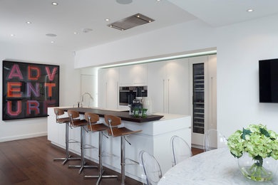 Large contemporary galley kitchen/diner in London with a submerged sink, flat-panel cabinets, white cabinets, composite countertops, integrated appliances, dark hardwood flooring, an island, brown floors and white worktops.