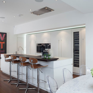 Large contemporary galley kitchen/diner in London with a submerged sink, flat-panel cabinets, white cabinets, composite countertops, integrated appliances, dark hardwood flooring, an island, brown floors and white worktops.
