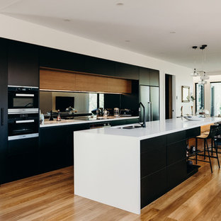 Inspiration for a contemporary open plan kitchen in Newcastle - Maitland with a double-bowl sink, flat-panel cabinets, black cabinets, mirror splashback, black appliances, with island, white benchtop and light hardwood floors.