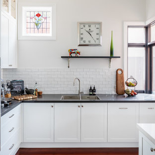 This is an example of a transitional l-shaped kitchen in Perth with shaker cabinets, white cabinets, quartz benchtops, white splashback, subway tile splashback, stainless steel appliances, with island, a double-bowl sink, medium hardwood floors, brown floor and black benchtop.