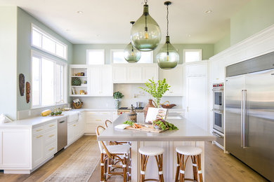 Example of a transitional u-shaped light wood floor enclosed kitchen design in Los Angeles with a farmhouse sink, shaker cabinets, white cabinets, white backsplash, stainless steel appliances, an island and white countertops