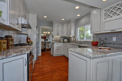 Mid-sized elegant l-shaped medium tone wood floor and brown floor open concept kitchen photo in San Francisco with an undermount sink, raised-panel cabinets, beige cabinets, granite countertops, blue backsplash, porcelain backsplash, stainless steel appliances, a peninsula and multicolored countertops