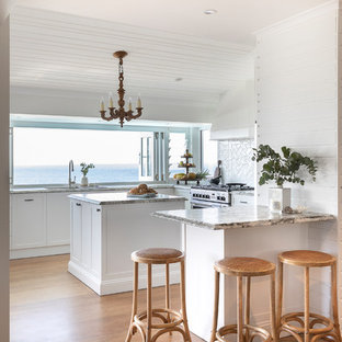Inspiration for a mid-sized tropical u-shaped kitchen in Sunshine Coast with an undermount sink, shaker cabinets, white cabinets, granite benchtops, white splashback, ceramic splashback, stainless steel appliances, medium hardwood floors, with island, multi-coloured benchtop and brown floor.