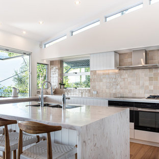 This is an example of a large beach style l-shaped eat-in kitchen in Sunshine Coast with a drop-in sink, white cabinets, beige splashback, porcelain splashback, black appliances, medium hardwood floors, with island and white benchtop.