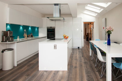 This is an example of a large contemporary l-shaped kitchen/diner in Surrey with a built-in sink, flat-panel cabinets, white cabinets, composite countertops, blue splashback, glass sheet splashback, stainless steel appliances, dark hardwood flooring, an island and white worktops.
