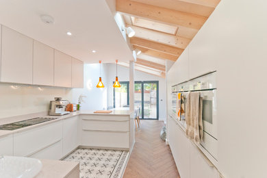 Photo of a contemporary u-shaped kitchen in Kent with flat-panel cabinets, white cabinets, white appliances, a breakfast bar, multi-coloured floors and white worktops.