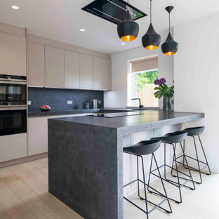 Design ideas for a medium sized contemporary galley kitchen in Cornwall with flat-panel cabinets, beige cabinets, black appliances, light hardwood flooring, a breakfast bar, beige floors and grey worktops.