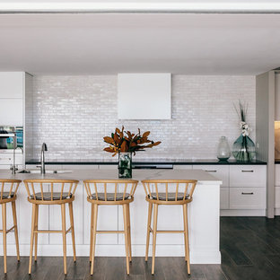 This is an example of a beach style galley kitchen in Sydney with an undermount sink, shaker cabinets, white cabinets, white splashback, panelled appliances, dark hardwood floors, with island, brown floor and black benchtop.
