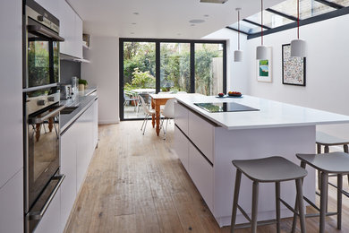 Inspiration for a medium sized contemporary kitchen/diner in London with white cabinets, an island, an integrated sink, flat-panel cabinets, composite countertops, grey splashback, stainless steel appliances and light hardwood flooring.