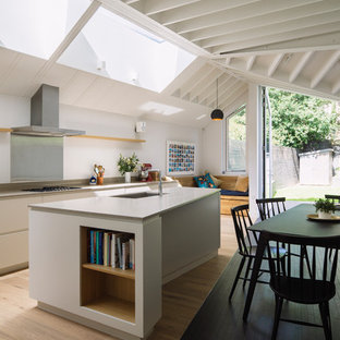 This is an example of a medium sized contemporary kitchen/diner in London with a built-in sink, flat-panel cabinets, white cabinets, composite countertops, grey splashback, light hardwood flooring, an island, brown floors and metal splashback.