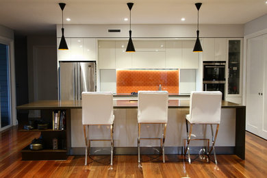Inspiration för mellanstora moderna kök, med en undermonterad diskho, släta luckor, vita skåp, bänkskiva i kvarts, orange stänkskydd, glaspanel som stänkskydd, rostfria vitvaror och en köksö