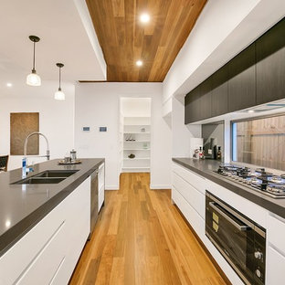 Photo of a contemporary galley open plan kitchen in Brisbane with a double-bowl sink, flat-panel cabinets, white cabinets, window splashback, black appliances, medium hardwood floors, with island, brown floor and grey benchtop.