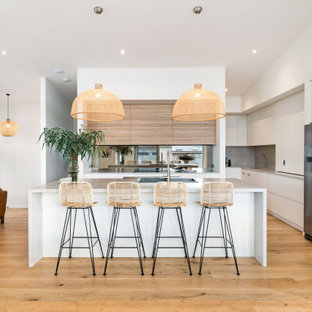 This is an example of a large beach style single-wall kitchen pantry in Wollongong with a double-bowl sink, flat-panel cabinets, white cabinets, metallic splashback, glass sheet splashback, stainless steel appliances, medium hardwood floors, with island, brown floor and beige benchtop.
