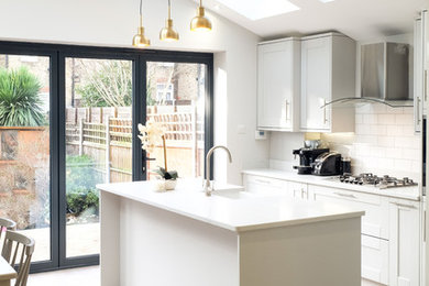 Design ideas for a medium sized classic single-wall open plan kitchen in Sussex with a belfast sink, shaker cabinets, grey cabinets, quartz worktops, white splashback, metro tiled splashback, stainless steel appliances, light hardwood flooring, an island, brown floors and white worktops.